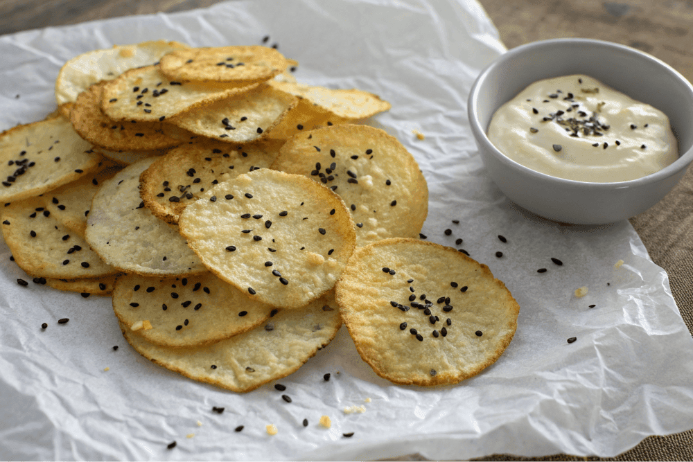 Homemade cottage cheese chips sprinkled with black sesame seeds, served with a creamy dipping sauce in a small bowl, arranged on parchment paper, rustic presentation, and healthy snack option.