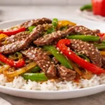 Pepper steak with sliced beef, red and green bell peppers, and onions in savory sauce served over white rice in a simple kitchen setting.